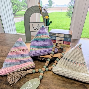 May include: Three crocheted hats in pastel shades, each with a pointed top. A wooden bead garland and a wooden sign that says "gather" are also present. A decorative arch with the word "hello" is in the background.