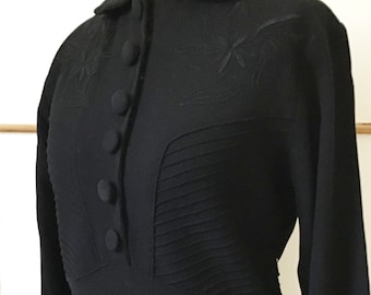 May include: Black long-sleeve blouse with a rounded collar and button-up front. The blouse features pleating details and embroidered floral designs on the shoulders. The vintage-style top is displayed on a mannequin.