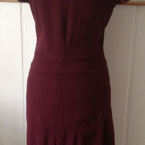 K&ouml;nnte beinhalten: Burgunderfarbenes Kleid mit kurzen &Auml;rmeln und knielangem Rock. Das Kleid wird auf einer Schaufensterpuppe pr&auml;sentiert, die seine ma&szlig;geschneiderte Passform und sein Design hervorhebt. Die R&uuml;ckseite des Kleides hat eine vertikale Naht und eine dezente Falte am Saum.