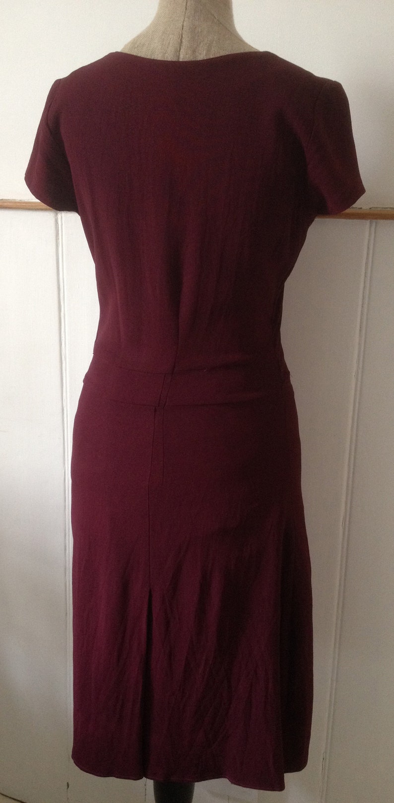 K&ouml;nnte beinhalten: Burgunderfarbenes Kleid mit kurzen &Auml;rmeln und knielangem Rock. Das Kleid wird auf einer Schaufensterpuppe pr&auml;sentiert, die seine ma&szlig;geschneiderte Passform und sein Design hervorhebt. Die R&uuml;ckseite des Kleides hat eine vertikale Naht und eine dezente Falte am Saum.