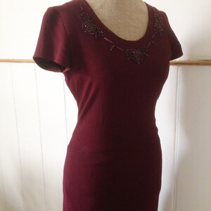 K&ouml;nnte beinhalten: Burgunderfarbenes Kleid mit kurzen &Auml;rmeln und einem Rundhalsausschnitt, verziert mit einem Perlendesign. Das Kleid wird auf einer Schaufensterpuppe pr&auml;sentiert.