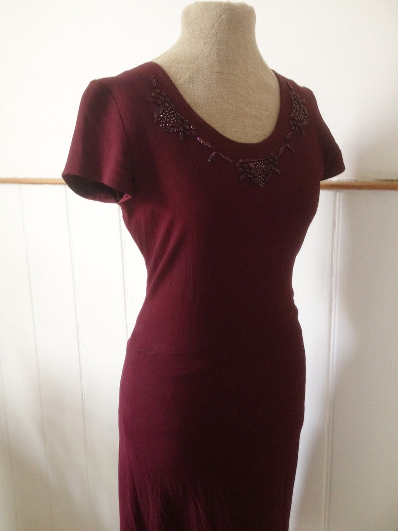 K&ouml;nnte beinhalten: Burgunderfarbenes Kleid mit kurzen &Auml;rmeln und einem Rundhalsausschnitt, verziert mit einem Perlendesign. Das Kleid wird auf einer Schaufensterpuppe pr&auml;sentiert.