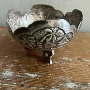 May include: A small, silver-colored bowl with a scalloped rim and three small feet. The bowl features an embossed floral design around the sides. The bowl is likely made of metal and sits on a wooden surface.