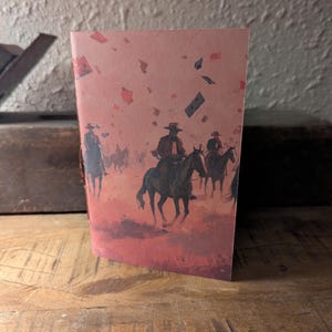 Może przedstawiać: Złożona karta z ilustracją w stylu western. Obraz przedstawia kowbojów na koniach na tle czerwonopomarańczowym z rozrzuconymi kształtami przypominającymi konfetti. Karta stoi na drewnianej powierzchni.