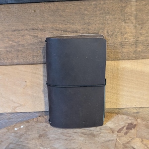 May include: A dark brown leather journal with a black elastic band closure. The journal is rectangular and appears to be closed. The background is a mix of light and dark brown wood.