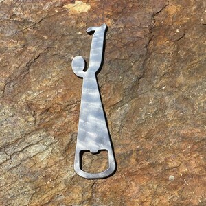 May include: A metal bottle opener in the shape of the letter "J". The opener has a silver finish with a textured appearance. The design includes a loop at the top and a bottle cap opener at the bottom. The background is a brown and tan rock surface.