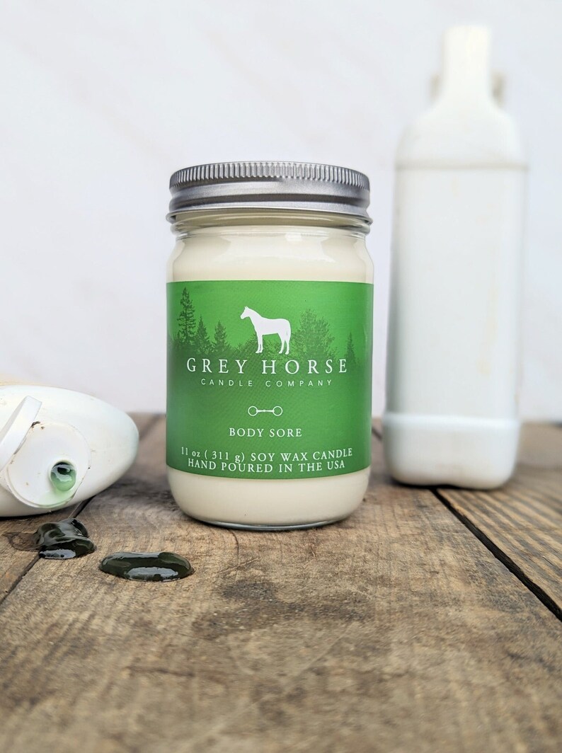 Puede incluir: Una vela de vidrio en un frasco con una etiqueta verde que dice "GREY HORSE CANDLE COMPANY" y "BODY SORE". La etiqueta tambi&eacute;n presenta una silueta blanca de un caballo y una herradura. La vela pesa 11 onzas (311 gramos) y est&aacute; hecha de cera de soya. Est&aacute; hecha a mano en los Estados Unidos.