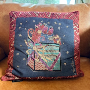 May include: Decorative square pillow with a navy blue background featuring a colorful illustration of cats in a gift box. The pillow has a red border with geometric patterns and star accents.