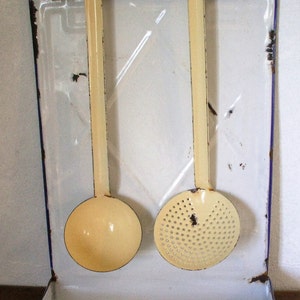 May include: White enamelware kitchen utensil holder with a blue rim. The holder has two hooks and a small shelf. Two yellow kitchen utensils are hanging from the hooks: a ladle and a slotted spoon.