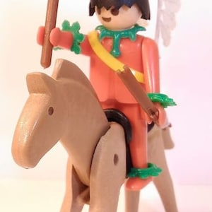 Op de afbeelding: Een plastic speelgoedfiguur van een indiaan die op een bruin paard rijdt. De figuur draagt een rode en gele outfit met een witte verenhoed. De figuur houdt een bruine speer vast met een groen bladontwerp.