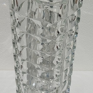 Peut inclure: Un vase en verre transparent avec un motif géométrique. Le vase est haut et de forme rectangulaire. Il est fait de verre épais et a une surface texturée.
