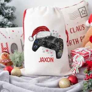 May include: A white canvas gift bag with a black and white video game controller wearing a red Santa hat. The bag has the name "Jaxon" printed on it.