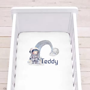 Può includere: Culla bianca con un lenzuolo con angoli bianchi con un'illustrazione ad acquerello di un astronauta in piedi sulla luna con un arcobaleno sopra. Il lenzuolo è personalizzato con il nome "Teddy".