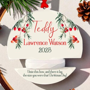 May include: A cream-coloured Christmas ornament with the name "Teddy" and "Lawrence Watson 2025" in red script. The ornament features a floral design and a red ribbon. The text "Untie this bow, and there it lay the size you were that Christmas Day!" is also included.