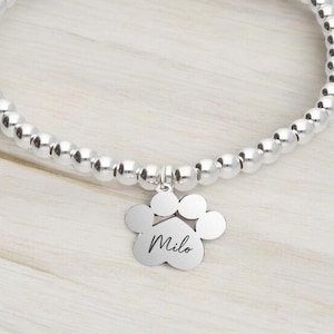 Pode incluir: Pulseira de prata com contas e um pingente de prata em forma de pata gravada com o nome "Milo".