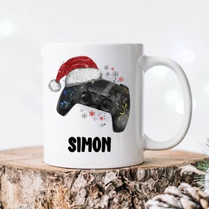以下が含まれることがあります： 赤いサンタの帽子をかぶった黒と緑のビデオゲームコントローラーが描かれた白いセラミックマグカップ。マグカップには白い雪の結晶と「サイモン」という名前が印刷されています。