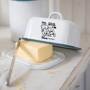 Op de afbeelding: Witte emaille boterdoos met een blauwe rand, een handvat en een bijpassend bord. De schaal heeft een zwarte afbeelding van brood en boter met de tekst "Jess, you're the butter to my bread love Tom x". Een mes en een blok boter liggen op het bord.