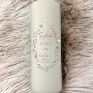 May include: A pale grey pillar candle decorated with a floral wreath. The wreath frames the name "Sophia", a cross, and the words "Christening Day" with a date and church name. The candle has a single wick.