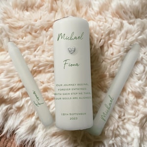 Puede incluir: Dos velas blancas con los nombres Michael y Fiona escritos en ellas. La vela más grande tiene el texto "Nuestro viaje comienza, para siempre entrelazados. Con cada paso que damos, nuestras almas están alineadas. 18 de septiembre de 2023".