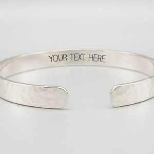 May include: A silver hammered cuff bracelet with a blank space for engraving. The text "YOUR TEXT HERE" is engraved on the bracelet.