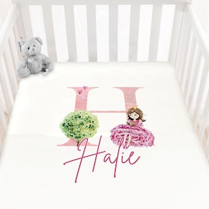Peut inclure: Un drap de lit blanc pour bébé avec une lettre H rose, une illustration de princesse et le nom "Hailie" écrit en cursive rose.