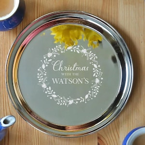 May include: A Christmas-themed table setting featuring a silver tray with the inscription "Christmas with the WATSON'S", surrounded by a floral wreath. Blue mugs, plates, a teapot, and yellow daffodils are also present.