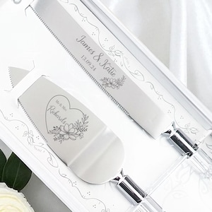 May include: A wedding cake knife and server set in a white box. The set includes a knife and server with clear acrylic handles and silver blades. The knife and server are engraved with names and a date. The box is decorated with floral designs.