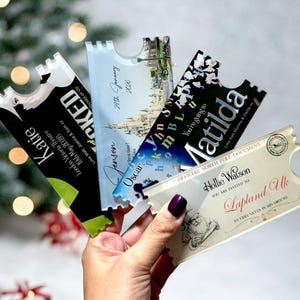 May include: A hand holds four clear acrylic tickets with various designs and text. One ticket reads "Katie" and "London Victoria Theatre." Another says "Jenson" and "29th January 2026." A third says "Matilda." The fourth says "Hollie Watson" and "Lapland Uk."
