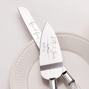 May include: A set of two silver cake knives with clear acrylic handles. The knives are engraved with the text "Katie & James" and "Mr. & Mrs." with the date "09.17.22".