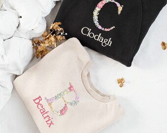 May include: Two cream colored sweatshirts with floral letter embroidery. The sweatshirt on the left has a pink "B" with the name "Beatrix" underneath. The sweatshirt on the right has a pink "C" with the name "Clodagh" underneath.