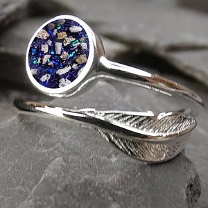 May include: A silver ring with a circular, dark blue centre containing small, colourful fragments. The ring's band has a feather design. The ring is set against a dark, textured background.
