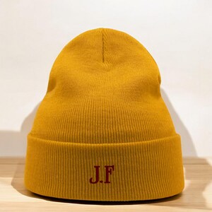 Puede incluir: Un gorro de lana amarillo mostaza con un "J.F." bordado en rojo en la parte delantera.