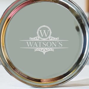Pode incluir: Uma bandeja redonda com borda prateada e borda interna dourada. O centro é verde suave com o nome "WATSON'S" e um monograma "W" branco. A bandeja está sobre uma superfície branca.