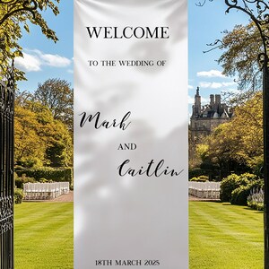 May include: A white wedding banner with black text that reads "WELCOME TO THE WEDDING OF Mark AND Caitlin 18TH MARCH 2025". The banner is displayed in a garden setting, with a castle visible in the distance.