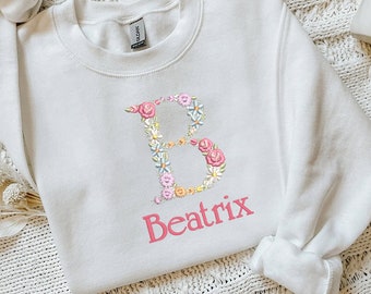 May include: White sweatshirt with a floral letter B and the name Beatrix printed in pink.