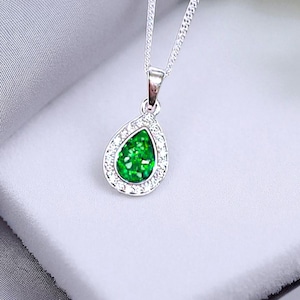 May include: A silver necklace with a teardrop pendant. The pendant has a bright green centre stone surrounded by a halo of clear, sparkling crystals. The necklace is displayed on a white surface.