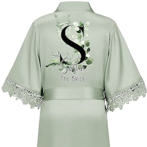 May include: Sage green satin robe with a lace trim. The robe features a large black letter "S" with green foliage and the text "Sasha The Bride".