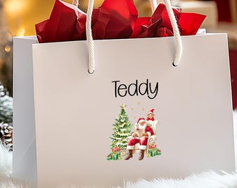 May include: A white gift bag with rope handles, filled with red tissue paper. The bag features the name "Teddy" in black script and a Christmas-themed illustration of Santa Claus and an elf. The bag is on a white, fluffy surface.