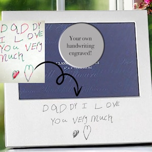 Puede incluir: Un marco de fotos blanco con un borde plateado. El marco tiene una abertura redonda en el centro donde se puede grabar un mensaje personalizado. El mensaje en el marco dice "DADDY I LOVE YOU VERY MUCH" con dos corazones dibujados debajo. El marco está sobre una mesa con un jarrón de flores rosas en el fondo.