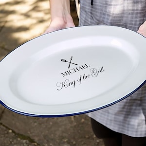 Op de afbeelding: Witte ovale serveerschaal met een blauwe rand. De schaal heeft een zwart ontwerp van gekruiste bestek en de tekst "MICHAEL King of the Grill".