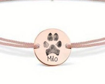 May include: A pink string bracelet with a rose gold charm featuring a paw print and the name "Milo" engraved on it.