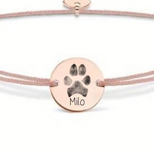 Può includere: Un braccialetto in corda rosa con un ciondolo in oro rosa che presenta un'impronta di zampa e il nome "Milo" inciso su di esso.