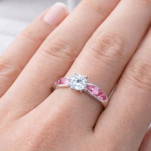 May include: A silver-toned ring featuring a central round-cut clear stone flanked by two marquise-shaped pink stones on each side of the band. The ring is displayed on a finger with a white jewellery box visible in the background.