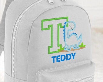 May include: A gray canvas backpack with a blue dinosaur and the letter T embroidered on the front. The name Teddy is embroidered below the dinosaur.