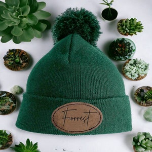 Pode incluir: Um gorro de malha verde com um remendo de couro marrom que diz "Forrest" em itálico. O gorro tem um pompom verde felpudo no topo.