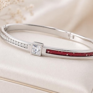 May include: A silver-toned hinged bangle bracelet featuring a row of small clear stones on one side, a central square-cut clear stone, and a deep red enamel inlay with subtle sparkling flecks on the opposite side of the metal band.