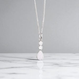 May include: A silver necklace with a pendant featuring two round, clear stones above a pear-shaped, light pink stone. The chain is a delicate curb link style, and the pendant hangs from a small loop.