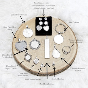 May include: A collection of silver-colored jewelry findings, including pendants, studs, a keychain, and a ring, arranged on a wooden slice. The items vary in shape and size, with some featuring ornate designs and others with clear settings.