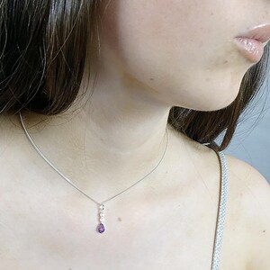 May include: A silver necklace with a delicate chain and a pendant featuring three small white stones and a teardrop-shaped purple gemstone. The necklace is worn around the neck.