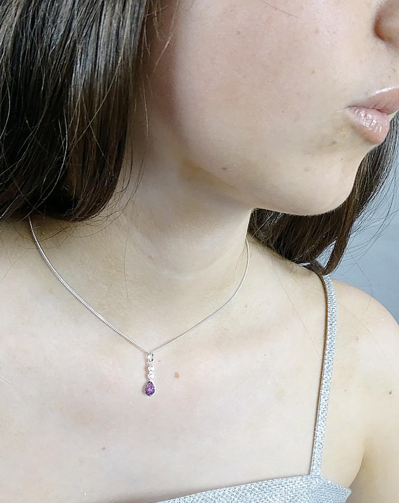 May include: A silver necklace with a delicate chain and a pendant featuring three small white stones and a teardrop-shaped purple gemstone. The necklace is worn around the neck.